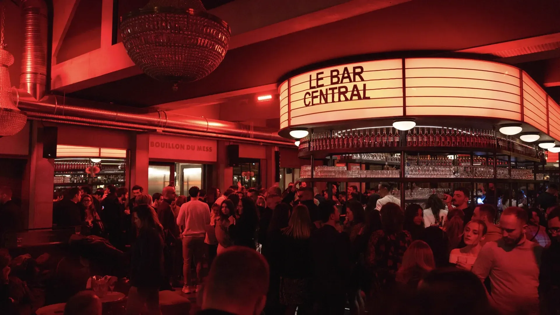 Foule dense dans un bar éclairé de rouge, autour d’un comptoir circulaire surmonté de l’enseigne « Le Bar Central »