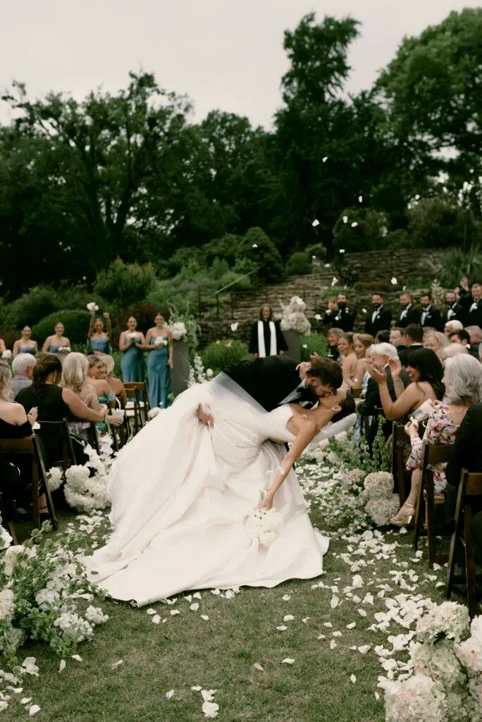 Dans l’allée d’une cérémonie en plein air, le marié renverse la mariée pour un baiser sous une pluie de pétales blancs.