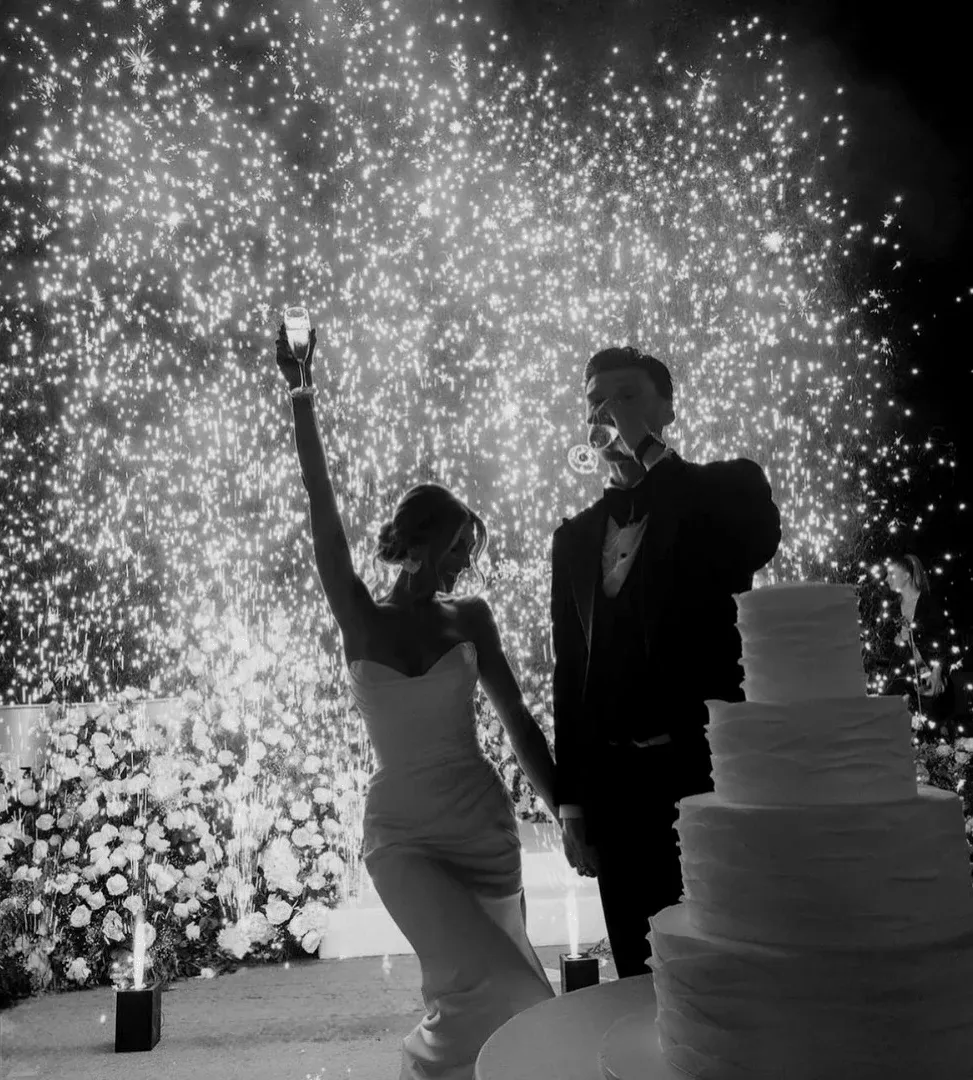 Les mariés portent un toast devant des étincelles froides, une pièce montée au premier plan. Photo en noir et blanc.