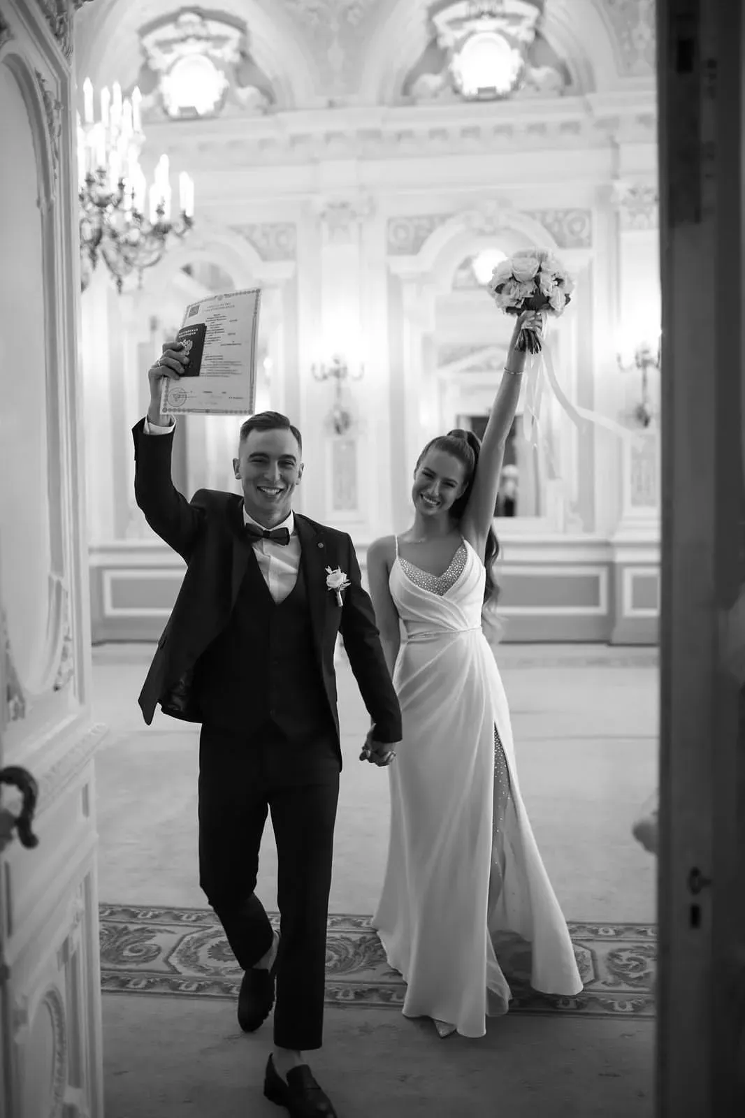 Les mariés avancent bras levés dans une grande salle aux moulures blanches : lui brandit un document, elle un bouquet blanc. Photo en noir et blanc.